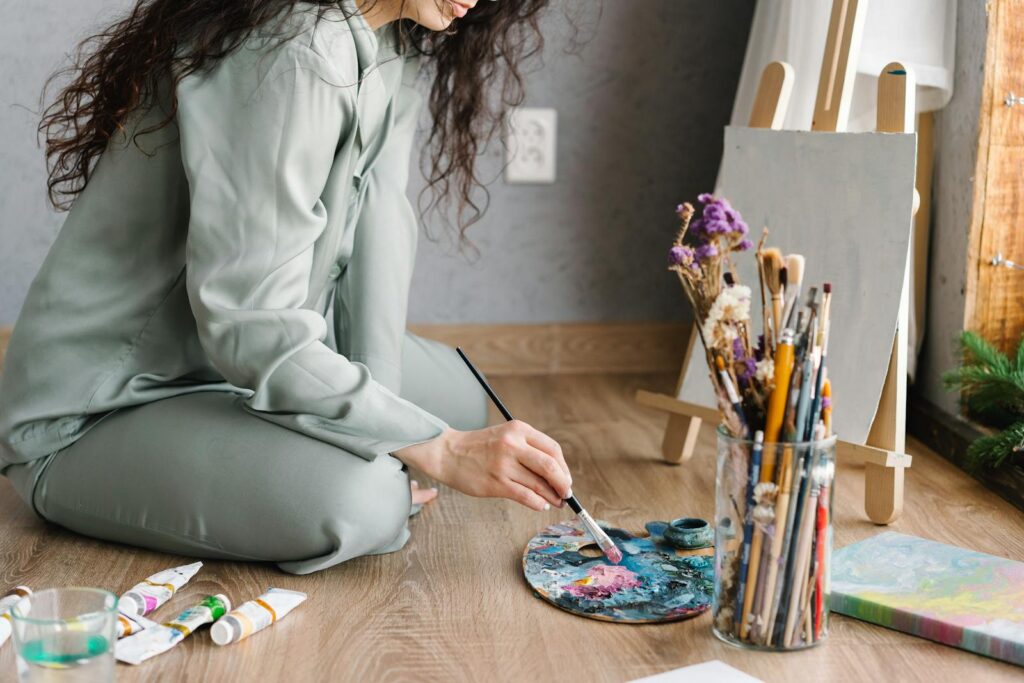 A woman artist in comfortable attire painting indoors with bright natural lighting.